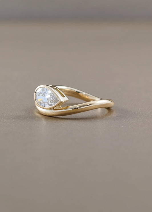 A gold ring with a pear-cut clear gemstone in a prong setting, resting on a smooth, warm gray surface.