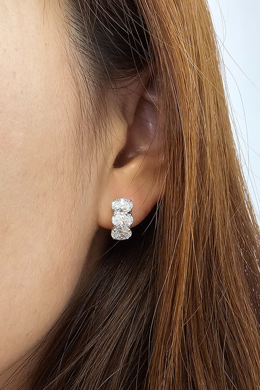 A close-up of a person's ear wearing a small silver stud earring with three diamond, against a light background.
