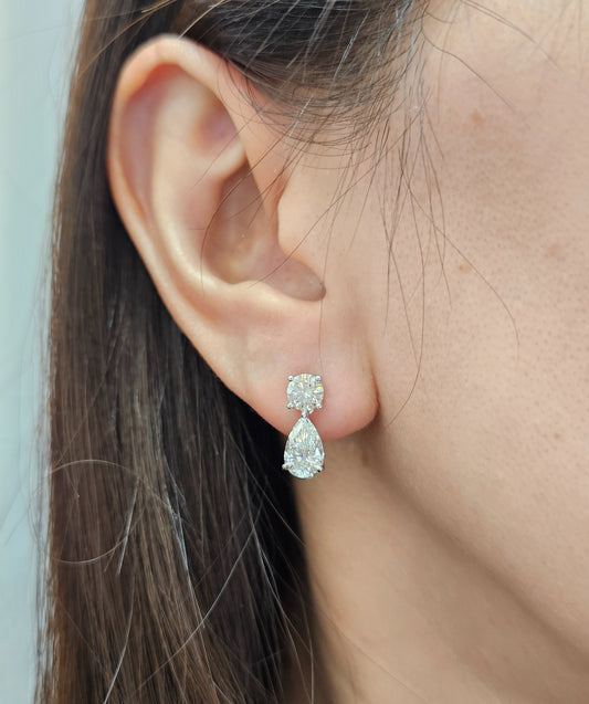 A close-up of a person's ear wearing a white stud earring with a small round clear gemstone above a larger pear-shaped clear gemstone.
