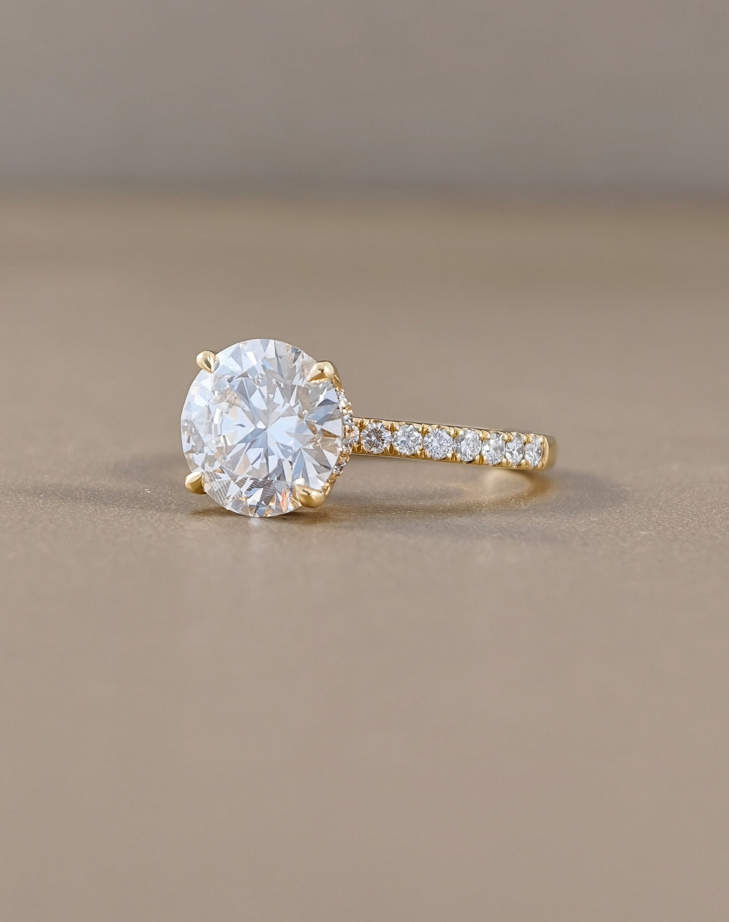 A yellow gold solitaire engagement ring with a round brilliant-cut clear center stone and a pavé-set diamond band, resting on a neutral beige surface.