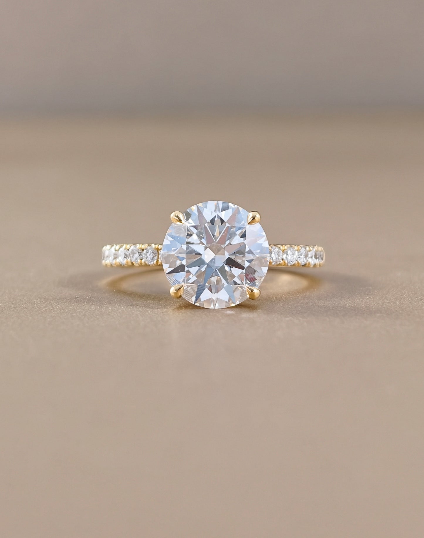 A yellow gold solitaire engagement ring featuring a round brilliant-cut clear center stone, with a pavé-set diamond band, photographed on a neutral beige background.