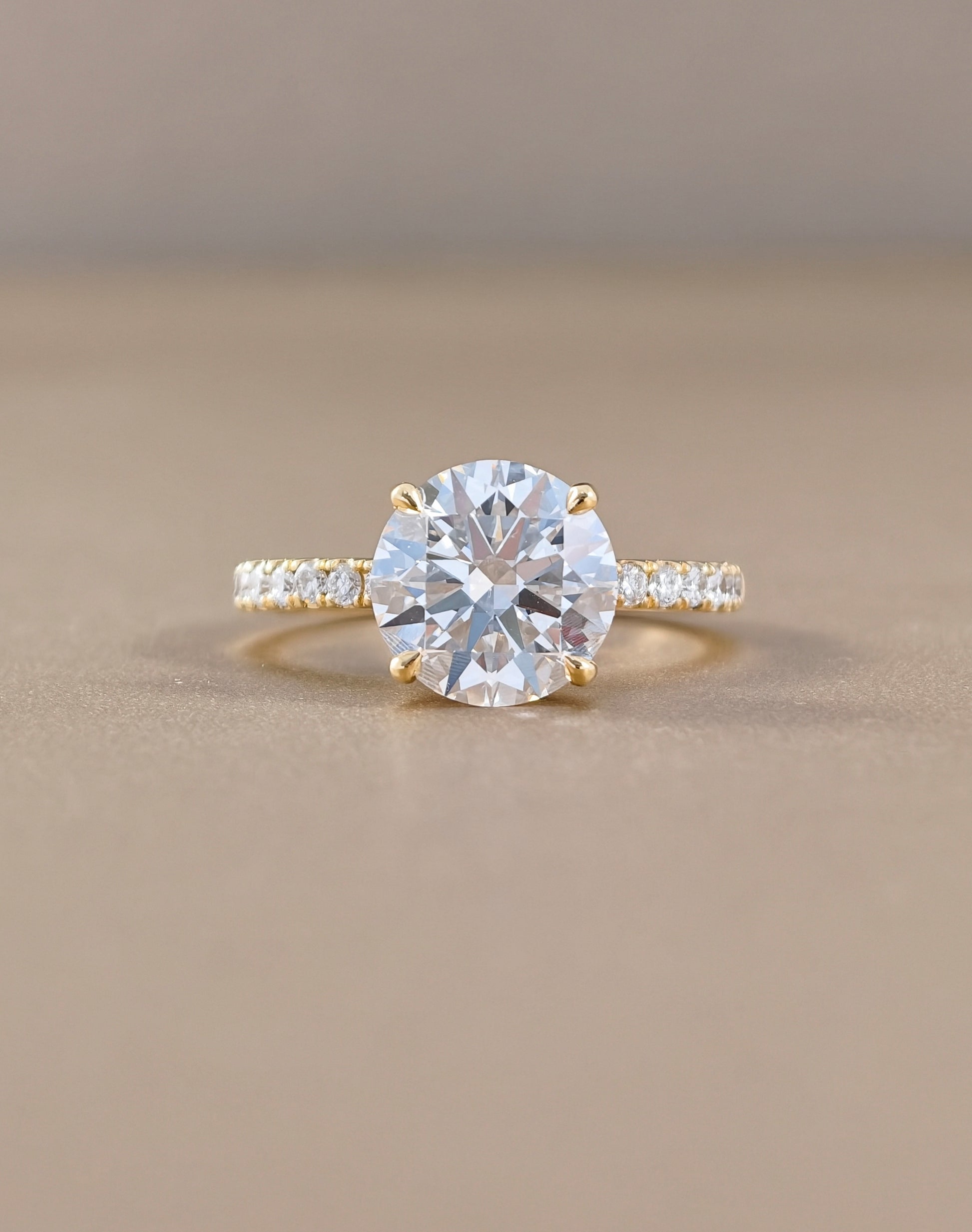 A yellow gold solitaire engagement ring featuring a round brilliant-cut clear center stone, with a pavé-set diamond band, photographed on a neutral beige background.
