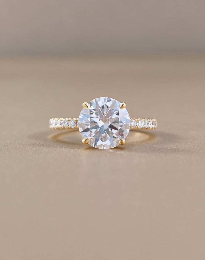 A yellow gold solitaire engagement ring featuring a round brilliant-cut clear center stone, with a pavé-set diamond band, photographed on a neutral beige background.