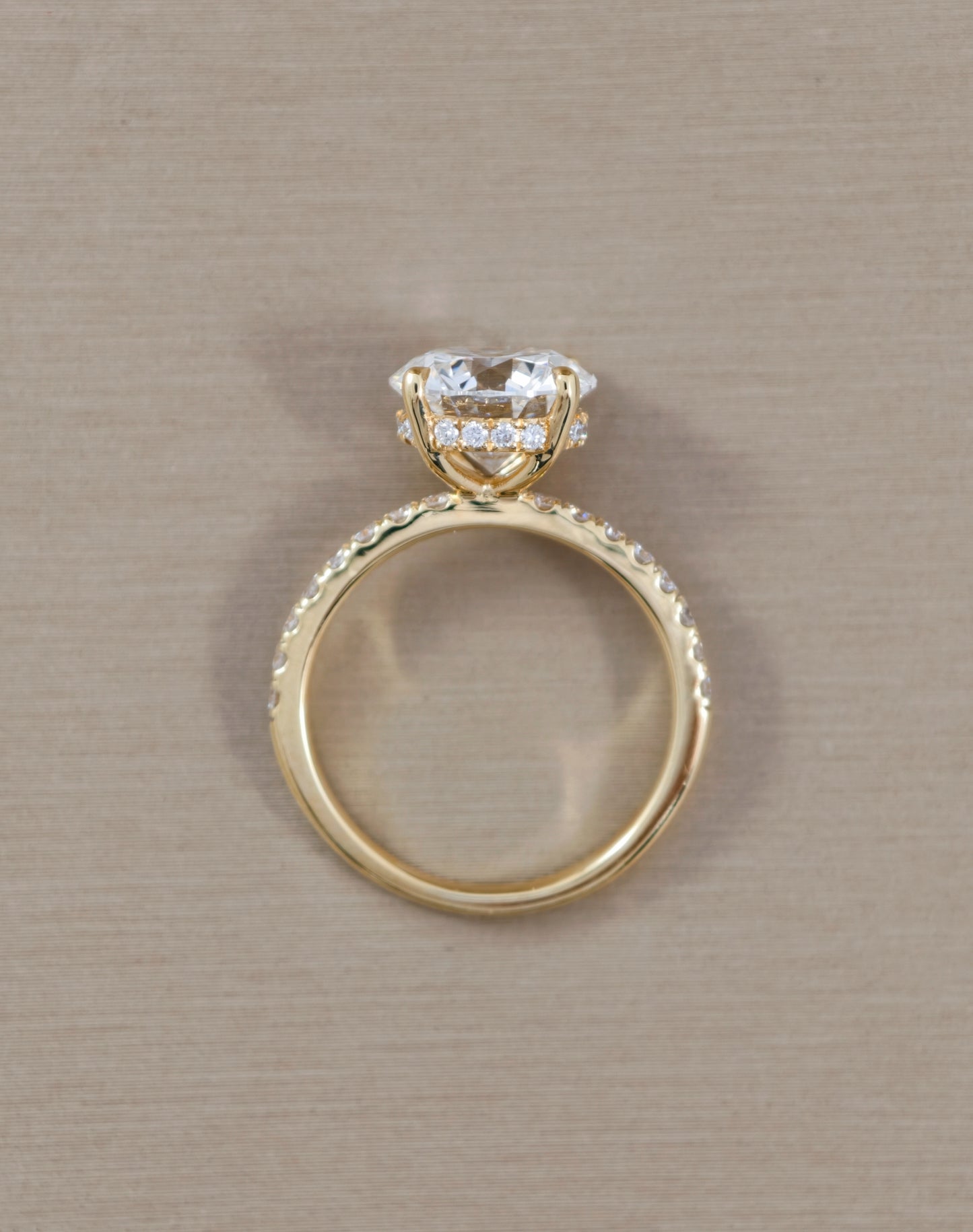 A yellow gold ring with a heart-shaped clear gemstone in a halo setting, accented with small pavé diamonds along the band, shown on a textured beige background.