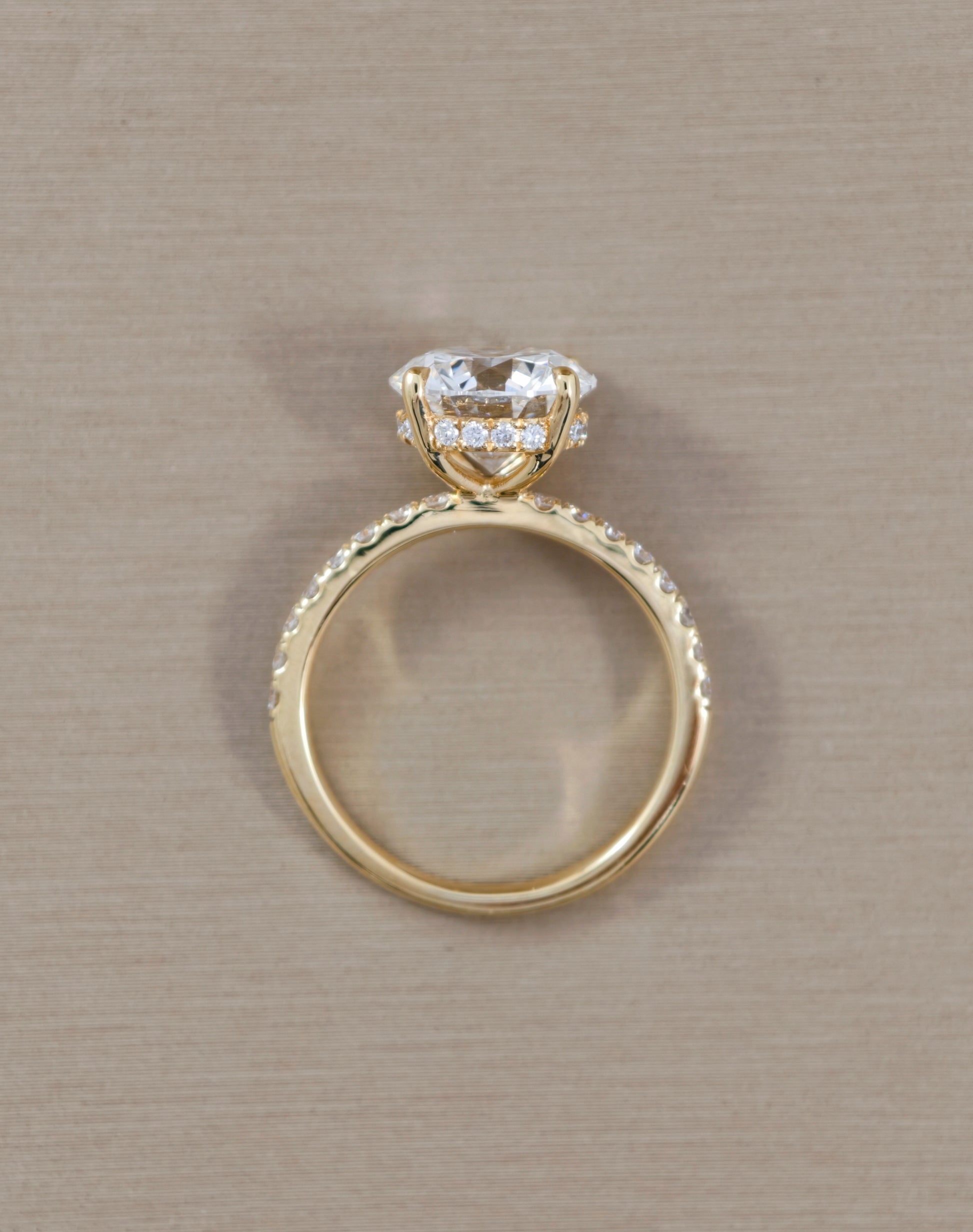 A yellow gold ring with a heart-shaped clear gemstone in a halo setting, accented with small pavé diamonds along the band, shown on a textured beige background.