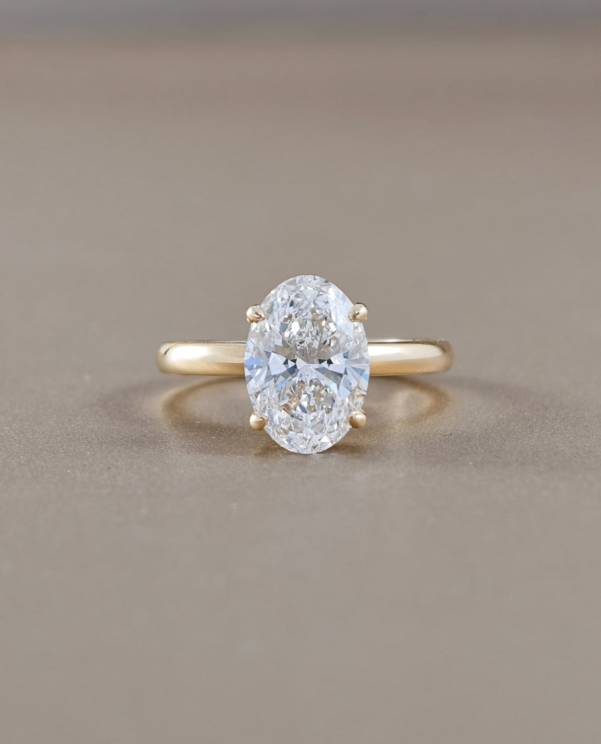 An oval-cut lab grown diamond ring set on a yellow gold band, photographed against a neutral, warm gray-brown background.