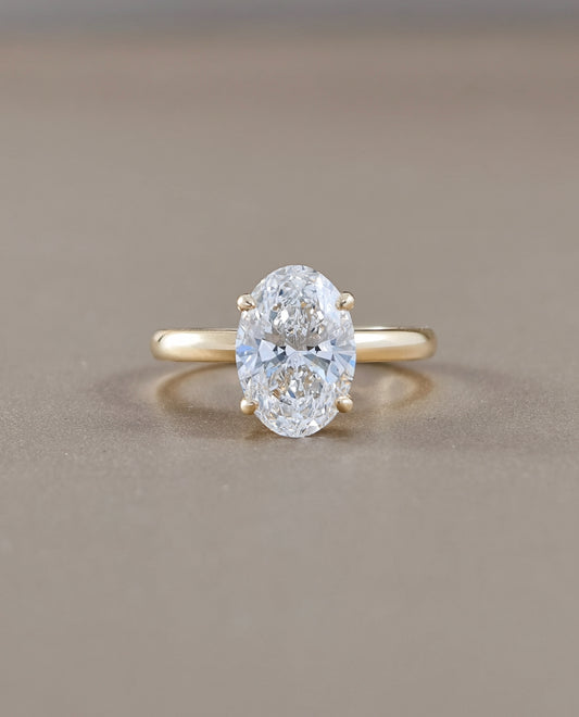 An oval-cut lab grown diamond ring set on a yellow gold band, photographed against a neutral, warm gray-brown background.