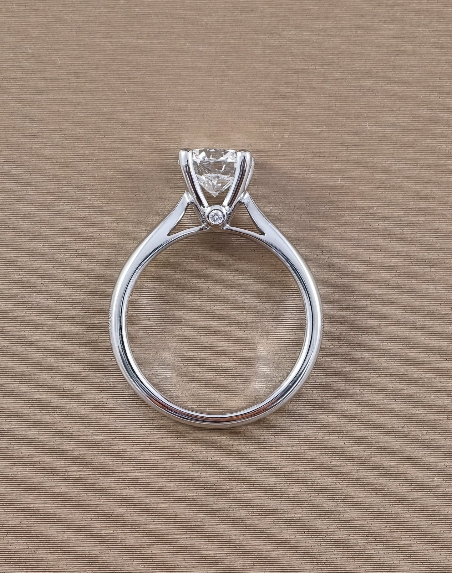 A white gold solitaire engagement ring with a round clear gemstone in a prong setting, featuring a small accent diamond on the band, shown on a textured beige background.