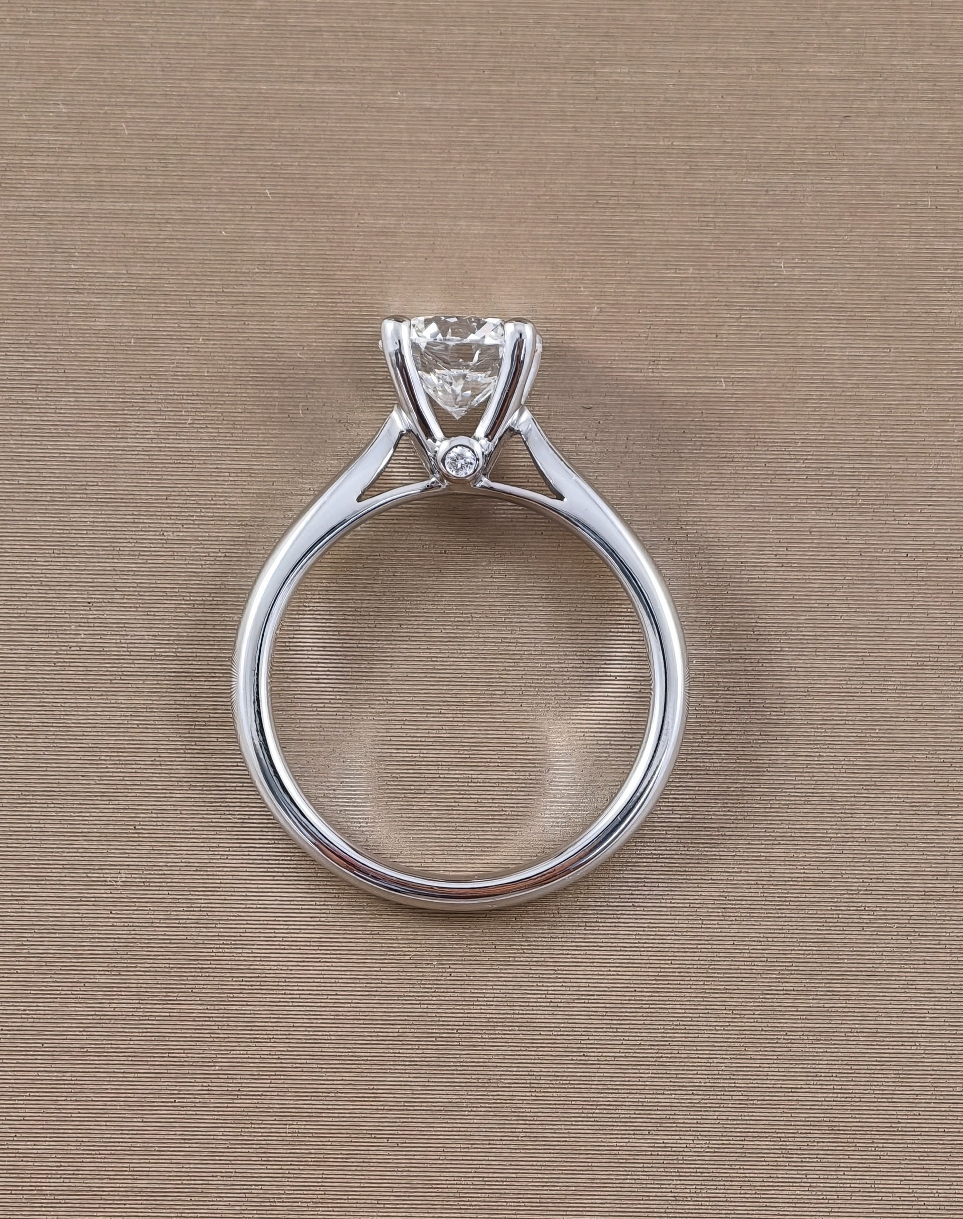 A white gold solitaire engagement ring with a round clear gemstone in a prong setting, featuring a small accent diamond on the band, shown on a textured beige background.