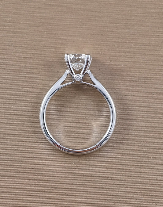 A white gold solitaire engagement ring with a round clear gemstone in a prong setting, featuring a small accent diamond on the band, shown on a textured beige background.
