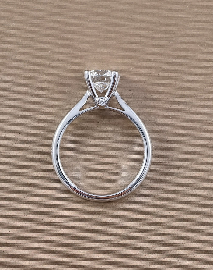 A white gold solitaire engagement ring with a round clear gemstone in a prong setting, featuring a small accent diamond on the band, shown on a textured beige background.