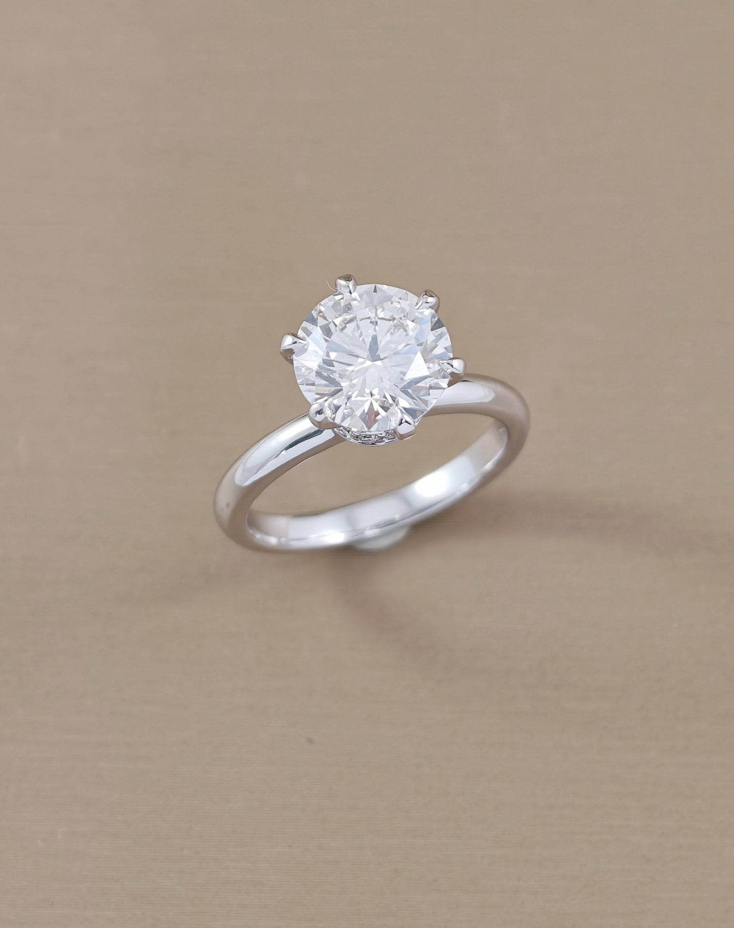 A classic solitaire engagement ring featuring a round brilliant-cut clear gemstone in a four-prong setting on a polished white gold, displayed on a textured beige background.