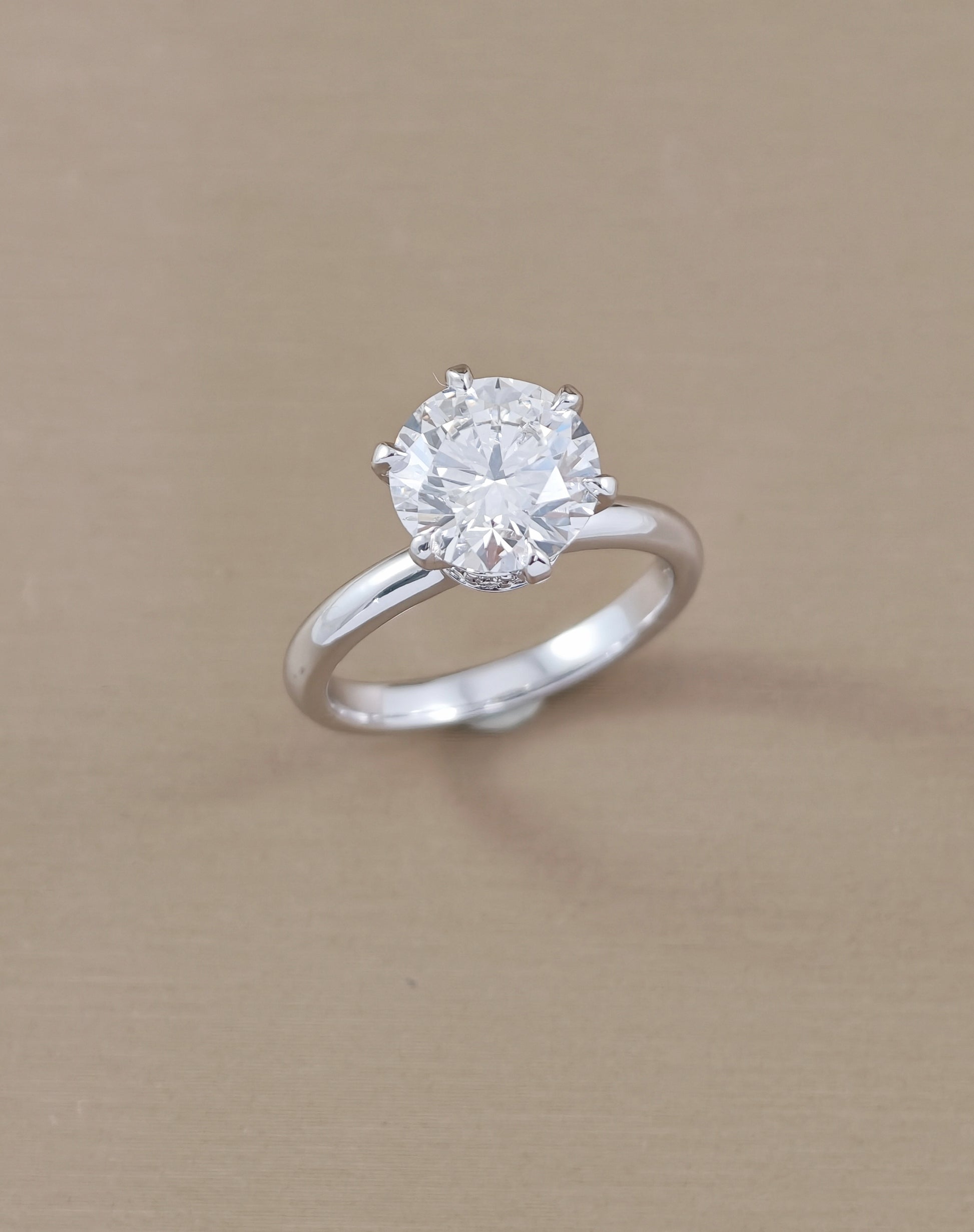 A classic solitaire engagement ring featuring a round brilliant-cut clear gemstone in a four-prong setting on a polished white gold, displayed on a textured beige background.