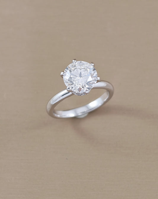 A classic solitaire engagement ring featuring a round brilliant-cut clear gemstone in a four-prong setting on a polished white gold, displayed on a textured beige background.