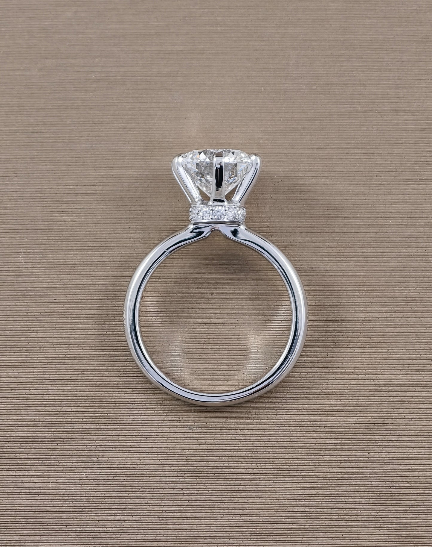 A white gold solitaire engagement ring with a round brilliant-cut diamond and a small pavé band of diamonds, shown on a textured beige background.