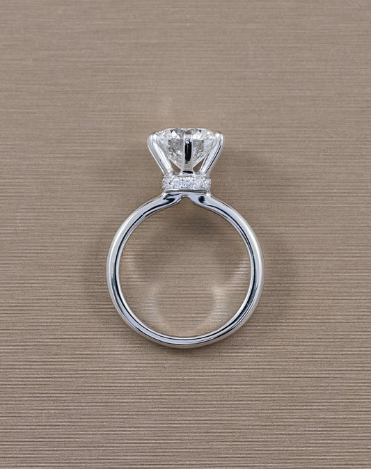 A white gold solitaire engagement ring with a round brilliant-cut diamond and a small pavé band of diamonds, shown on a textured beige background.