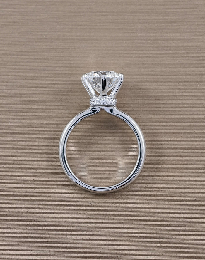 A white gold solitaire engagement ring with a round brilliant-cut diamond and a small pavé band of diamonds, shown on a textured beige background.