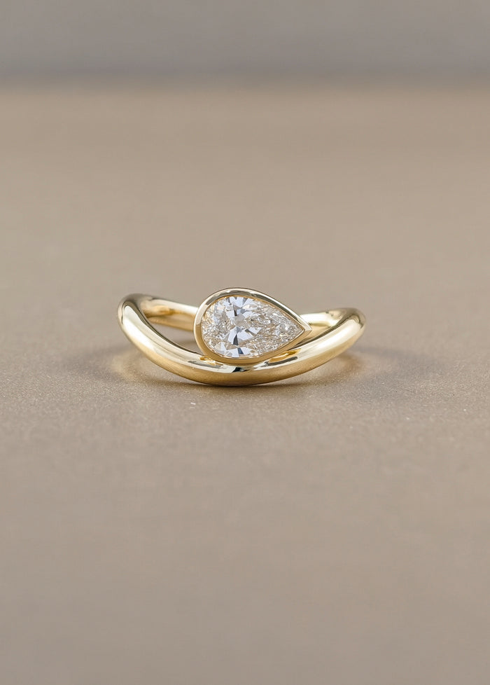 A gold ring featuring a pear-cut clear gemstone in a bezel setting, mounted on a curved, twisted gold band, photographed on a neutral beige background.