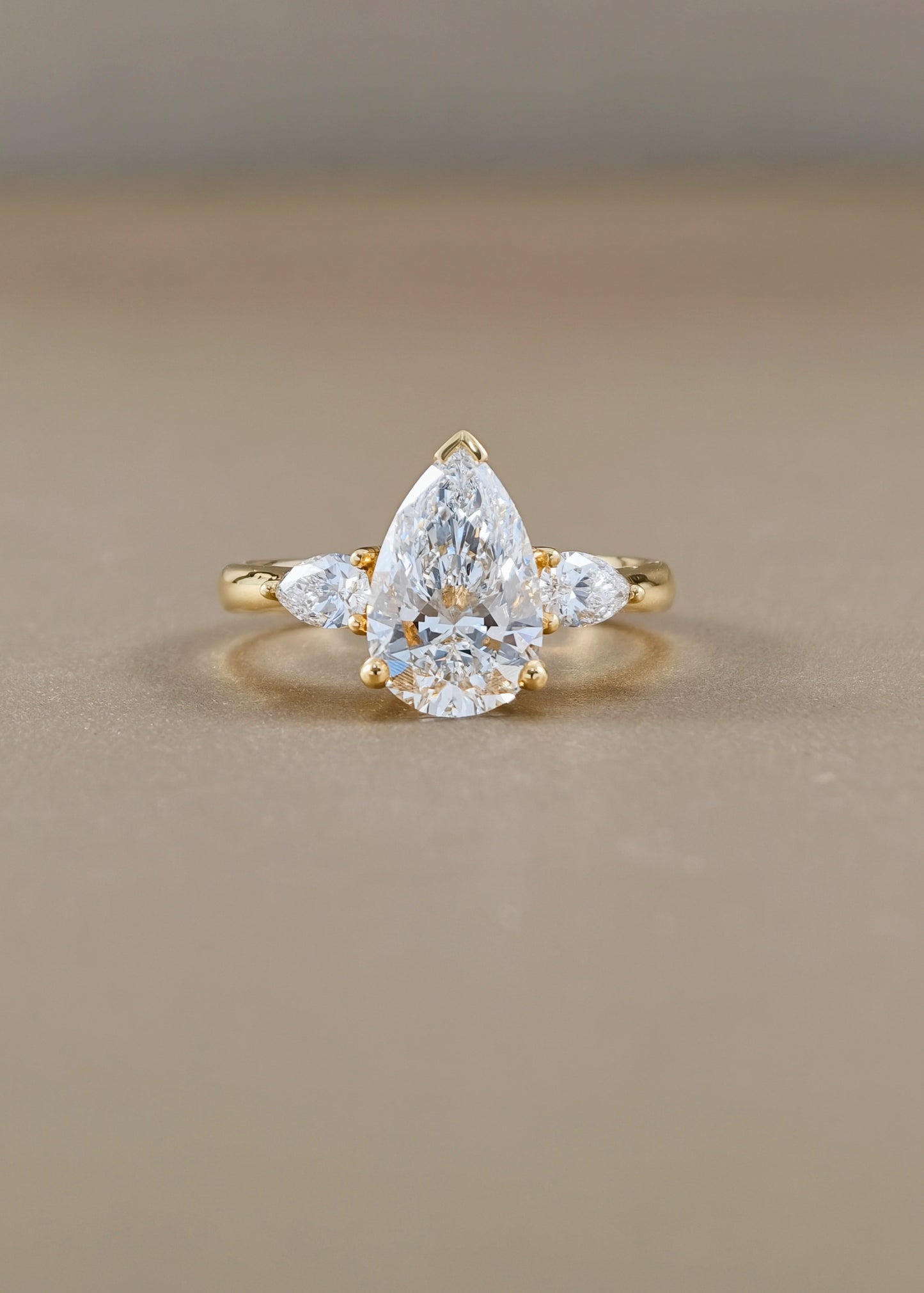A yellow gold ring featuring a pear-cut clear center stone with two smaller marquise-cut side stones, photographed on a smooth beige surface.