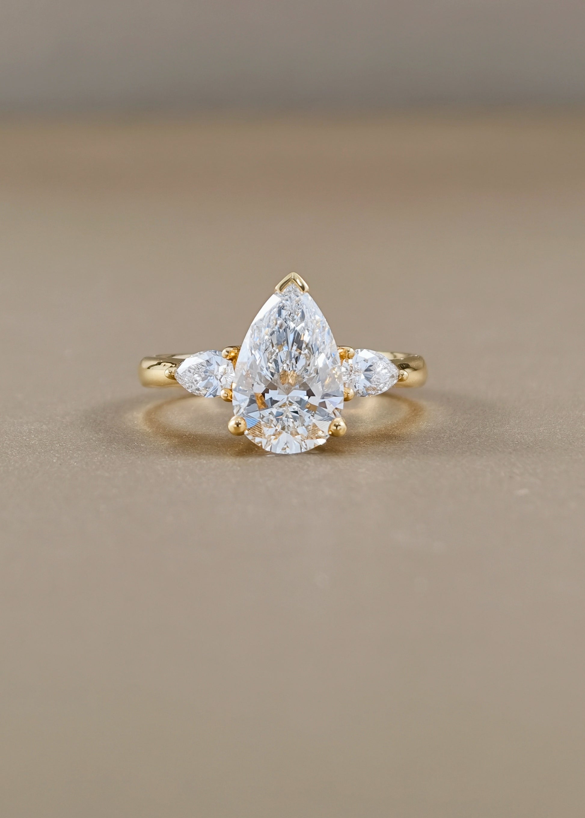 A yellow gold ring featuring a pear-cut clear center stone with two smaller marquise-cut side stones, photographed on a smooth beige surface.