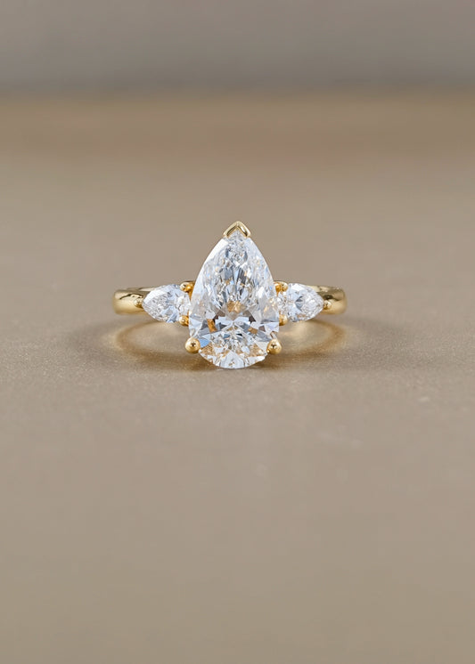 A yellow gold ring featuring a pear-cut clear center stone with two smaller marquise-cut side stones, photographed on a smooth beige surface.