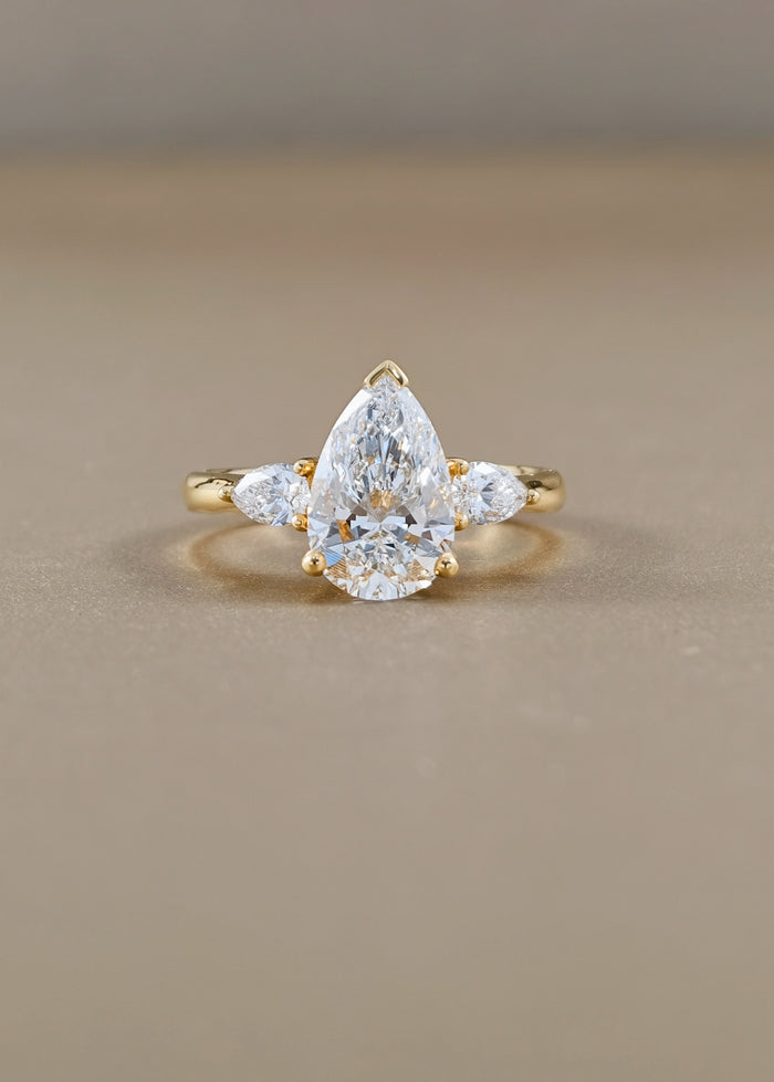 A yellow gold ring featuring a pear-cut clear center stone with two smaller marquise-cut side stones, photographed on a smooth beige surface.
