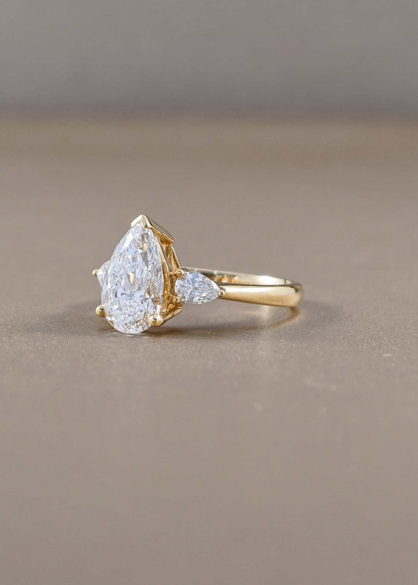 A gold ring featuring a pear-cut clear gemstone and a smaller marquise-cut clear gemstone on a warm taupe background.