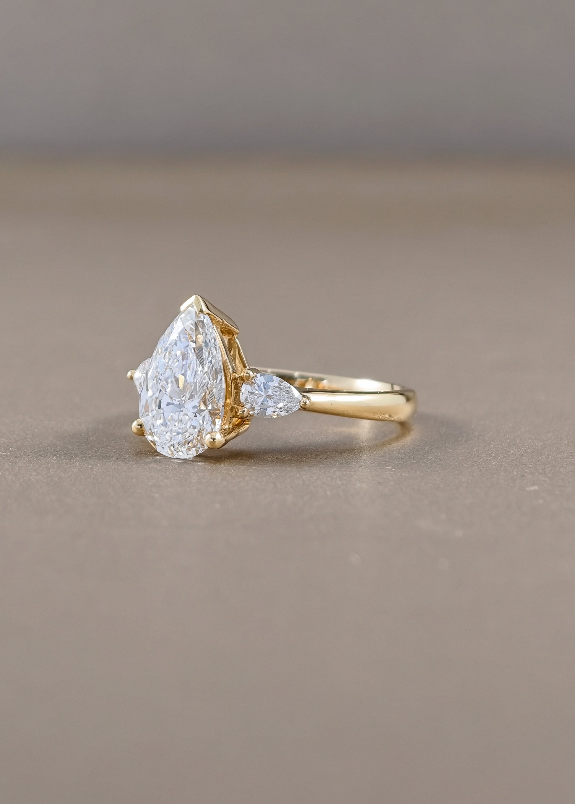 A gold ring featuring a pear-cut clear gemstone and a smaller marquise-cut clear gemstone on a warm taupe background.