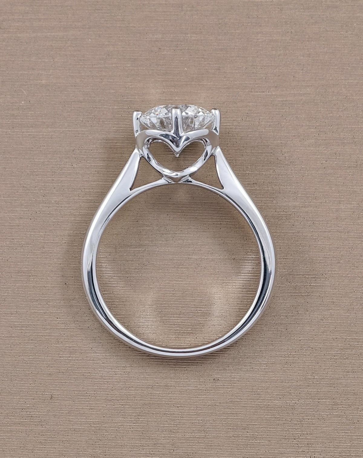 A white gold heart-shaped ring with a clear heart-shaped gemstone, photographed from a top-down perspective on a textured beige background.