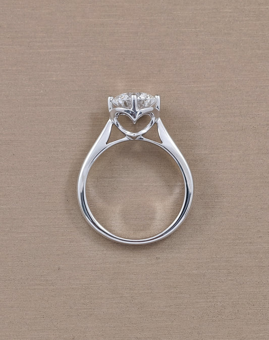 A white gold heart-shaped ring with a clear heart-shaped gemstone, photographed from a top-down perspective on a textured beige background.