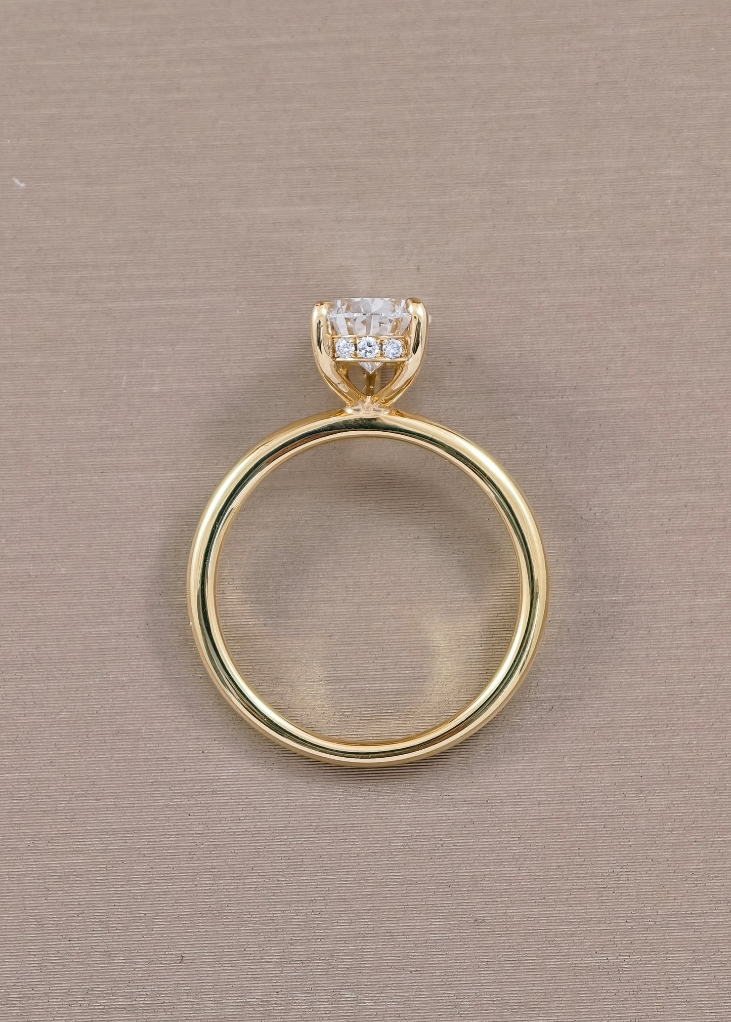 A yellow gold ring with a single round-cut clear gemstone in a prong setting, shown from the side against a textured beige background.