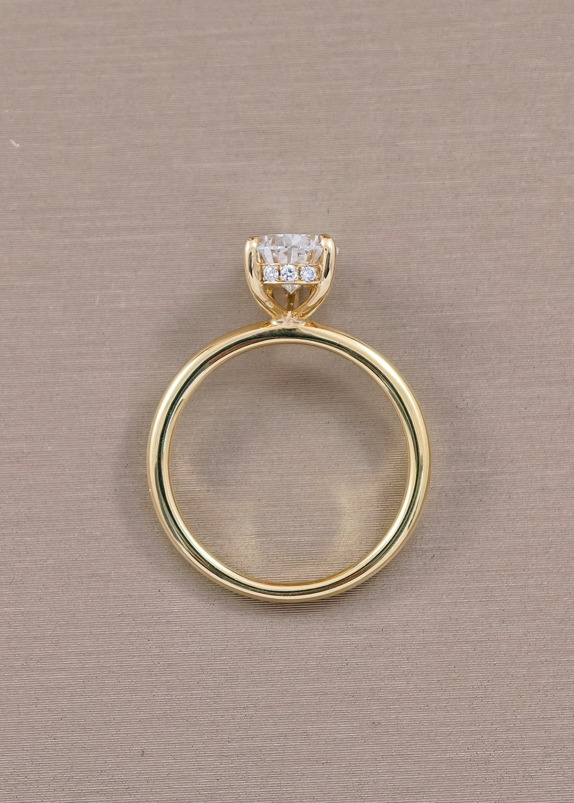 A yellow gold ring with a single round-cut clear gemstone in a prong setting, shown from the side against a textured beige background.
