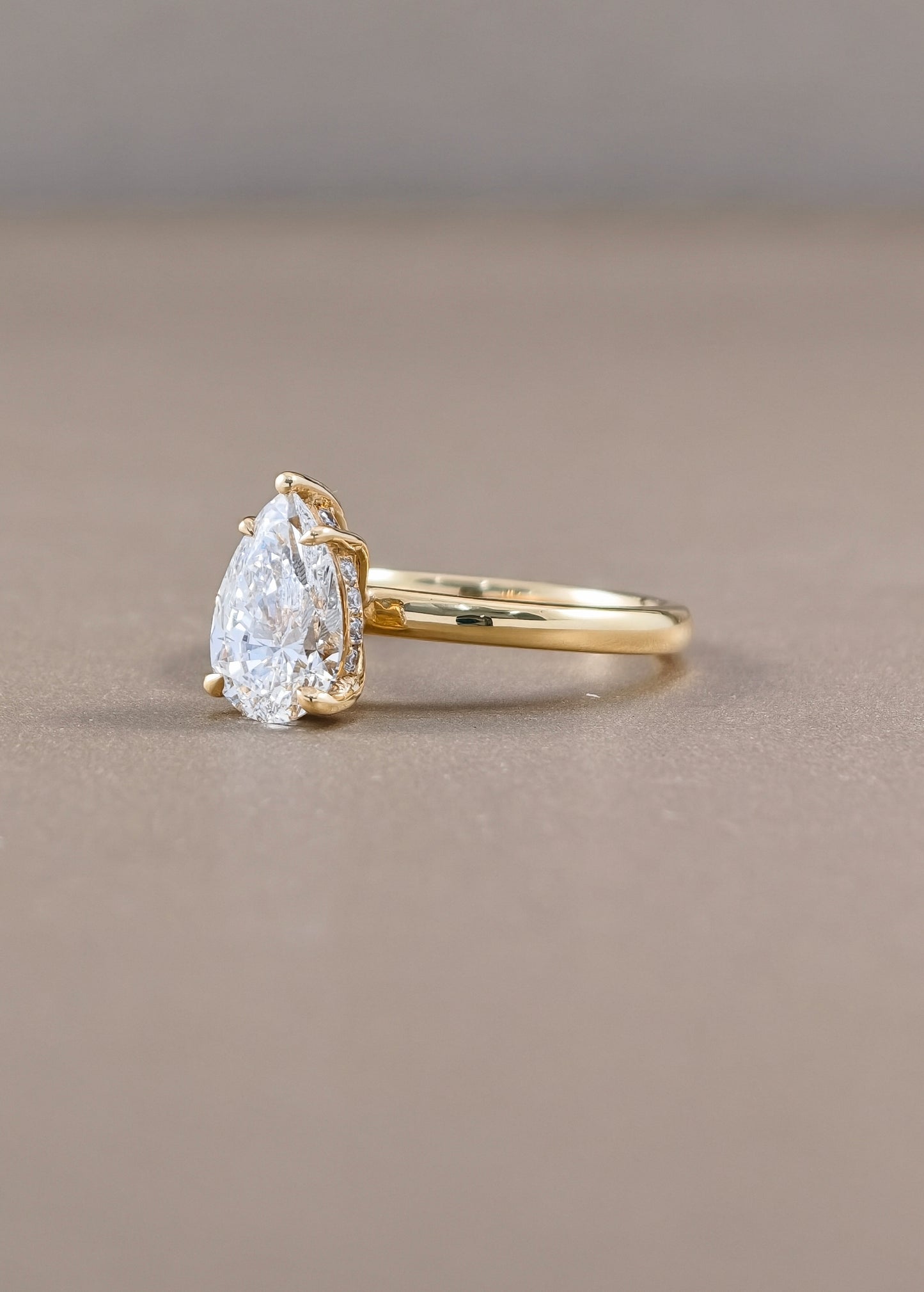 A pear-cut diamond ring with a yellow gold band, photographed on a neutral taupe background.