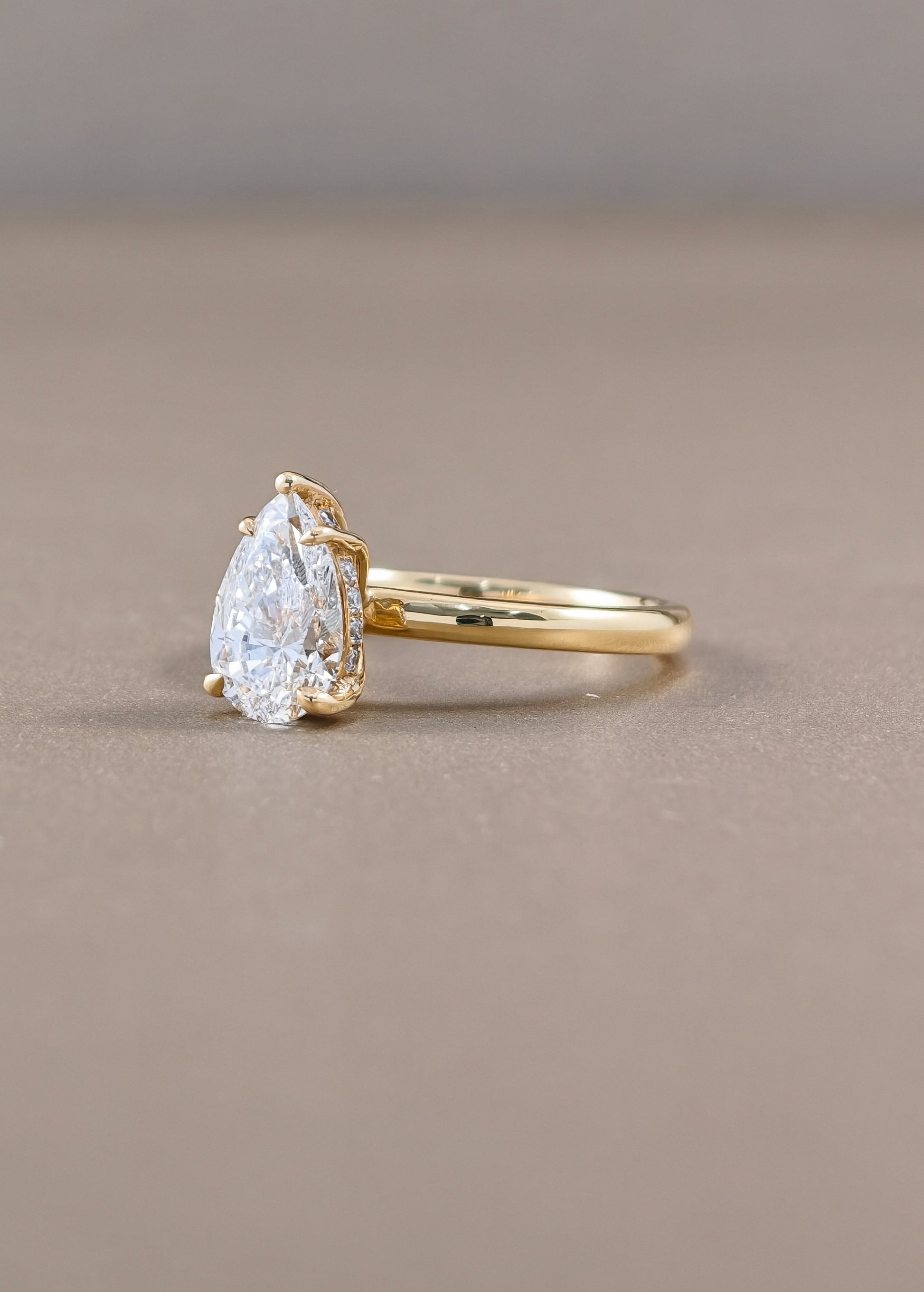A pear-cut diamond ring with a yellow gold band, photographed on a neutral taupe background.