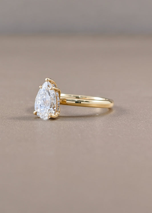 A pear-cut diamond ring with a yellow gold band, photographed on a neutral taupe background.
