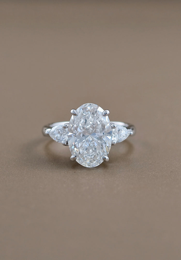 An oval diamond engagement ring with pear-cut side stones in a white gold-colored setting, photographed on a smooth, solid taupe-brown background.