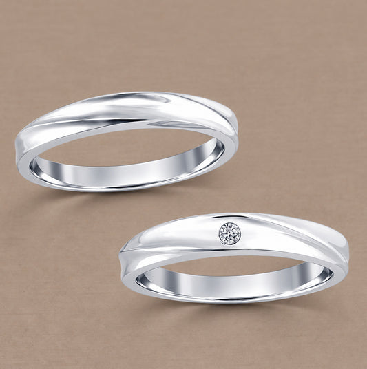 Two polished white gold  rings on a warm beige background, with the lower ring featuring a small round clear gemstone accent.