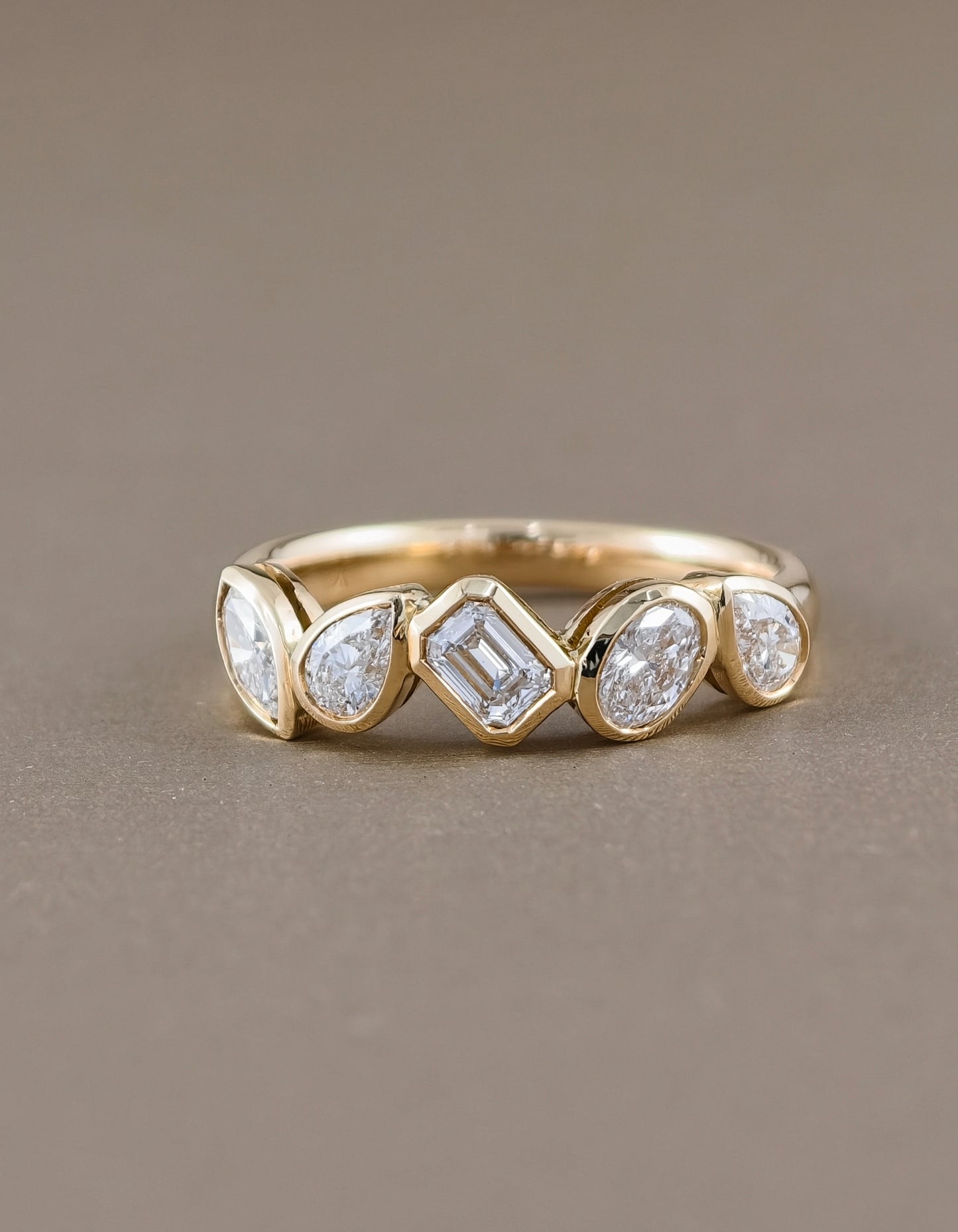 A gold band ring featuring five bezel-set clear gemstones in varied shapes, photographed on a smooth, solid taupe background.
