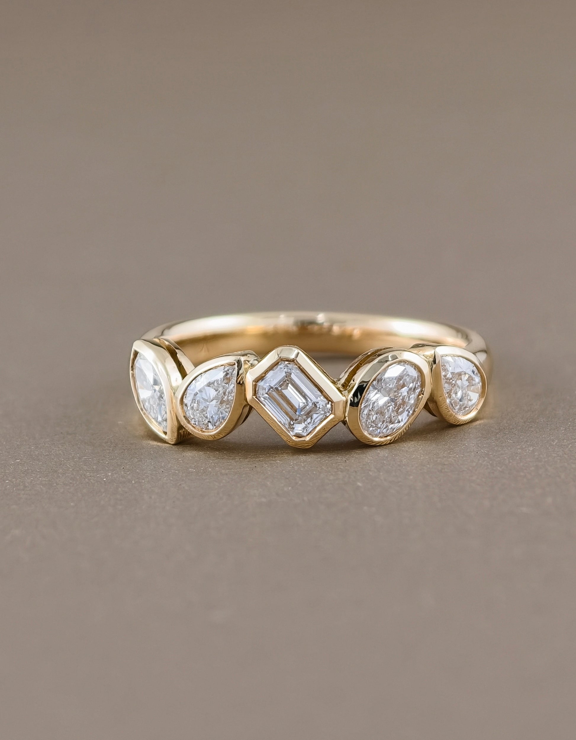 A gold band ring featuring five bezel-set clear gemstones in varied shapes, photographed on a smooth, solid taupe background.