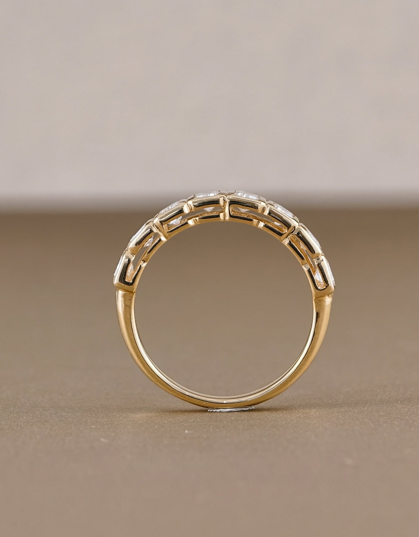 A gold ring with a decorative geometric pattern of small clear stones along the upper band, photographed on a neutral beige background.