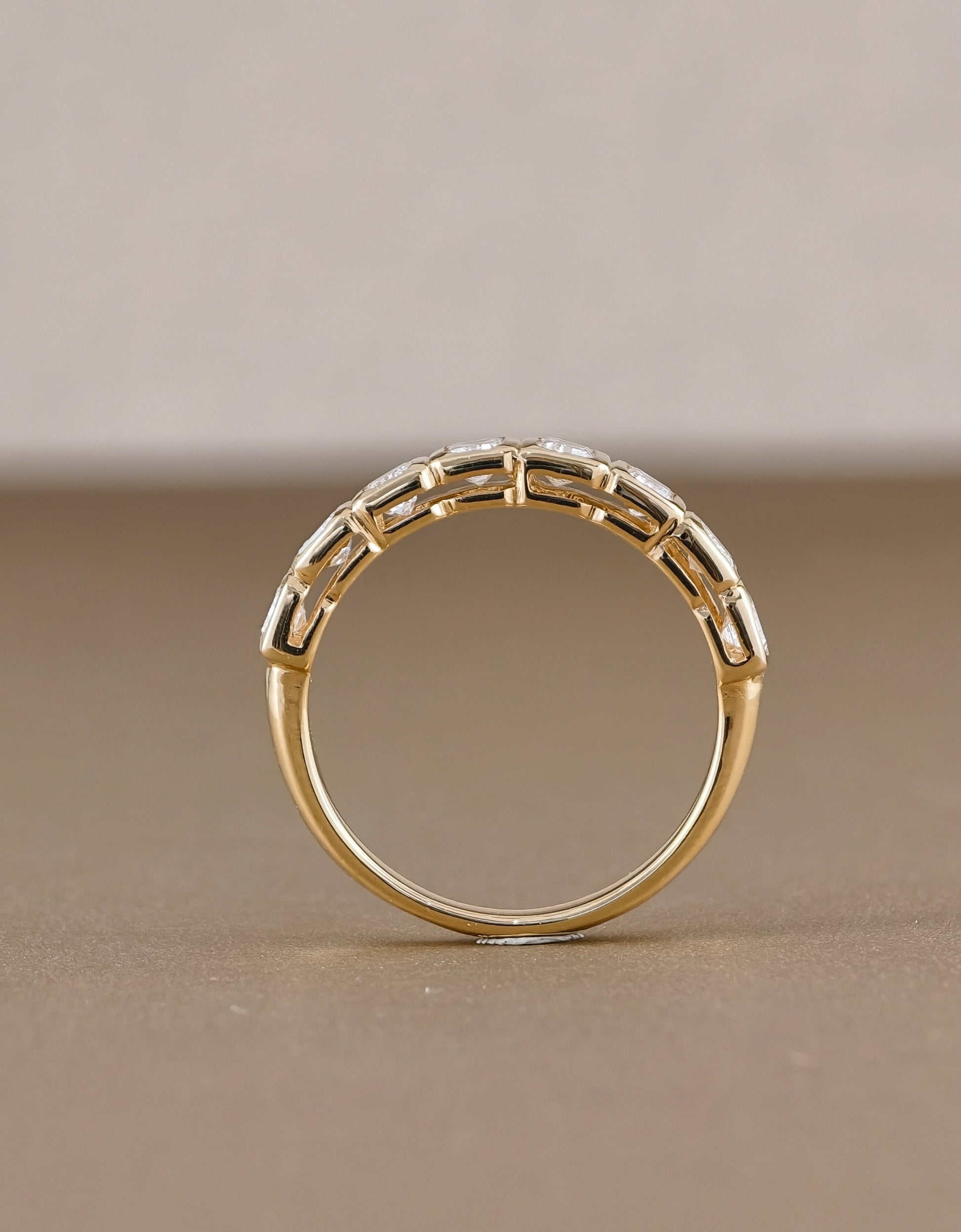 A gold ring with a decorative geometric pattern of small clear stones along the upper band, photographed on a neutral beige background.
