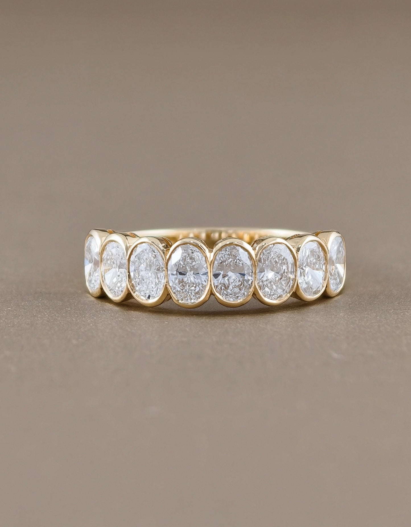 A gold eternity ring featuring seven oval-cut clear gemstones in a bezel setting, displayed on a neutral taupe background.