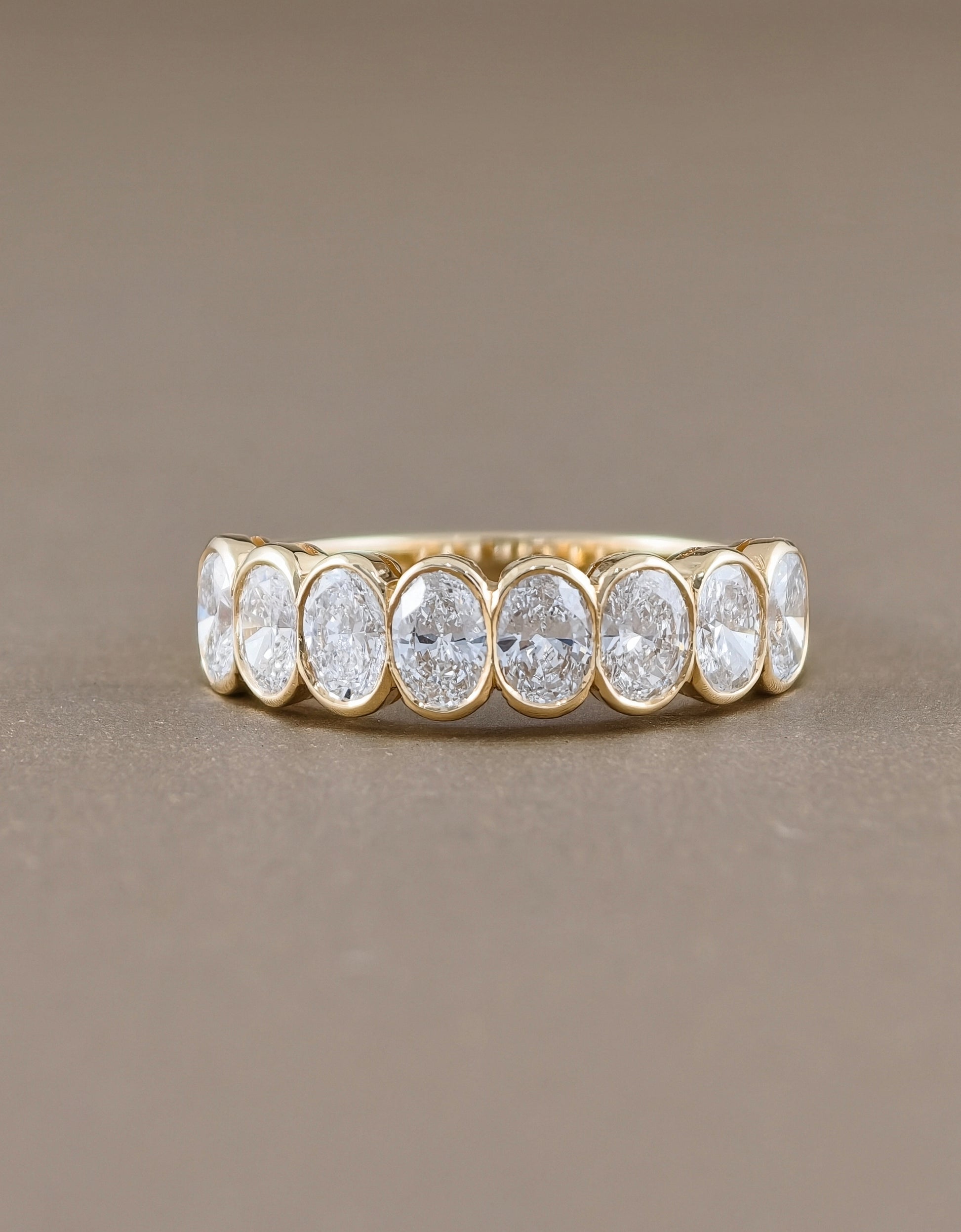 A gold eternity ring featuring seven oval-cut clear gemstones in a bezel setting, displayed on a neutral taupe background.