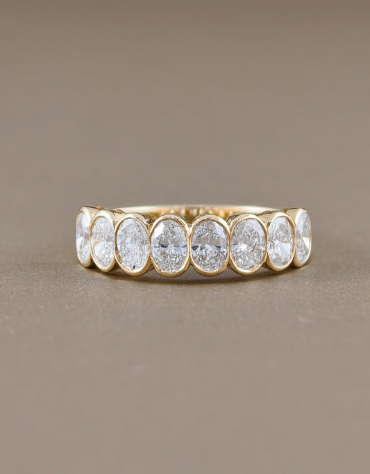 A gold eternity ring featuring seven oval-cut clear gemstones in a bezel setting, displayed on a neutral taupe background.