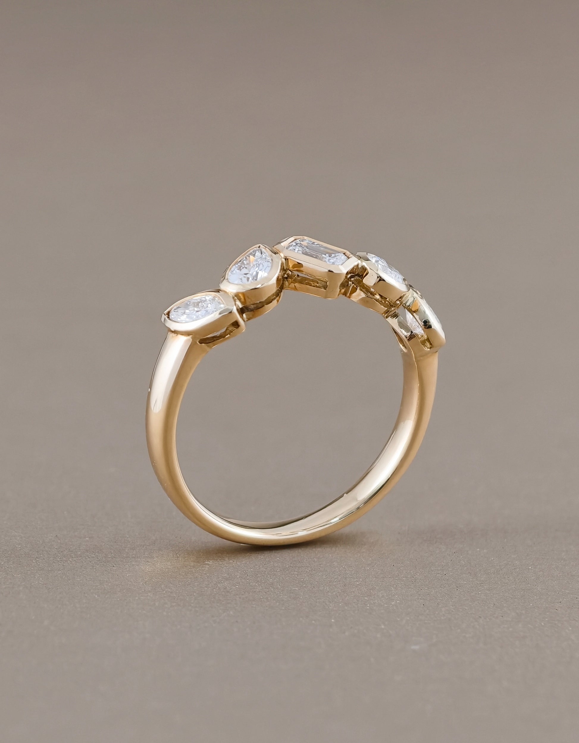 A yellow gold ring featuring a row of marquise-cut clear gemstones in a prong setting, displayed on a neutral taupe background.