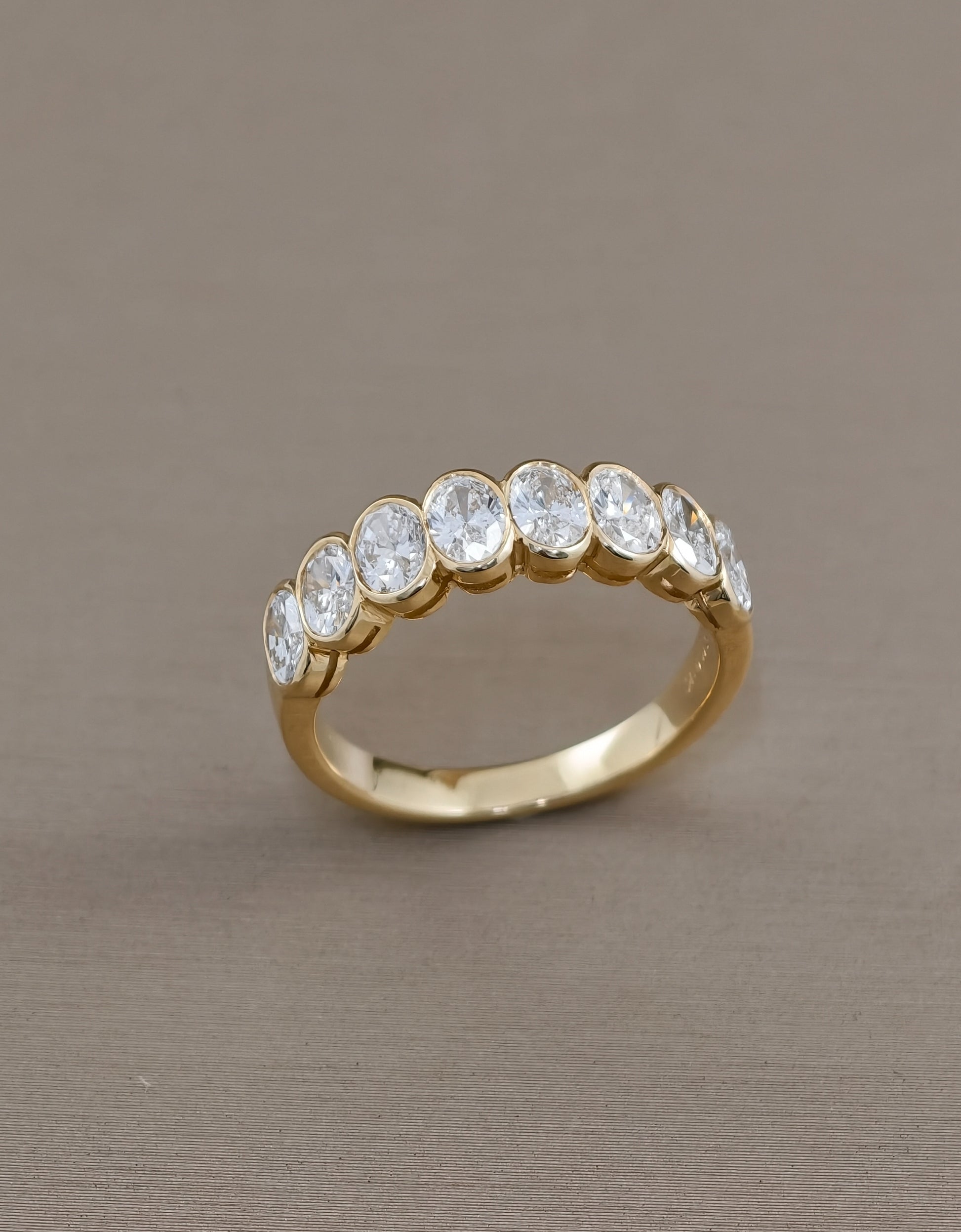 A gold eternity ring featuring a row of round-cut clear gemstones set in a polished yellow gold band, photographed on a neutral taupe background.