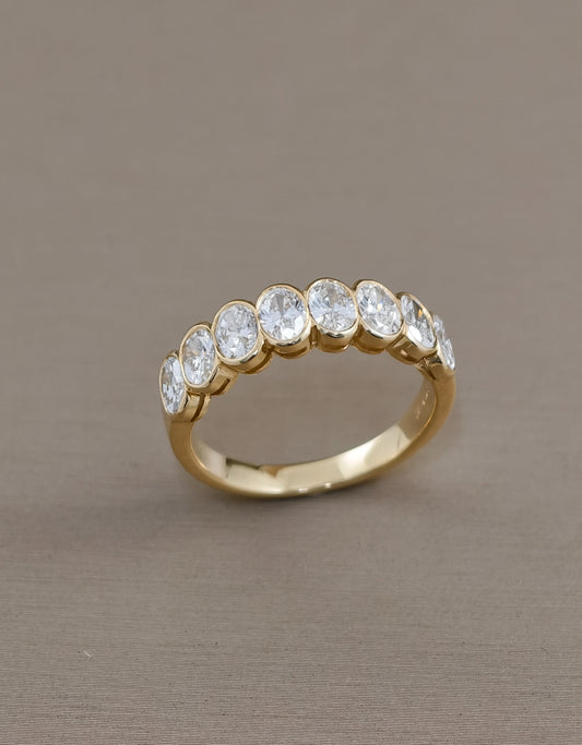A gold eternity ring featuring a row of round-cut clear gemstones set in a polished yellow gold band, photographed on a neutral taupe background.