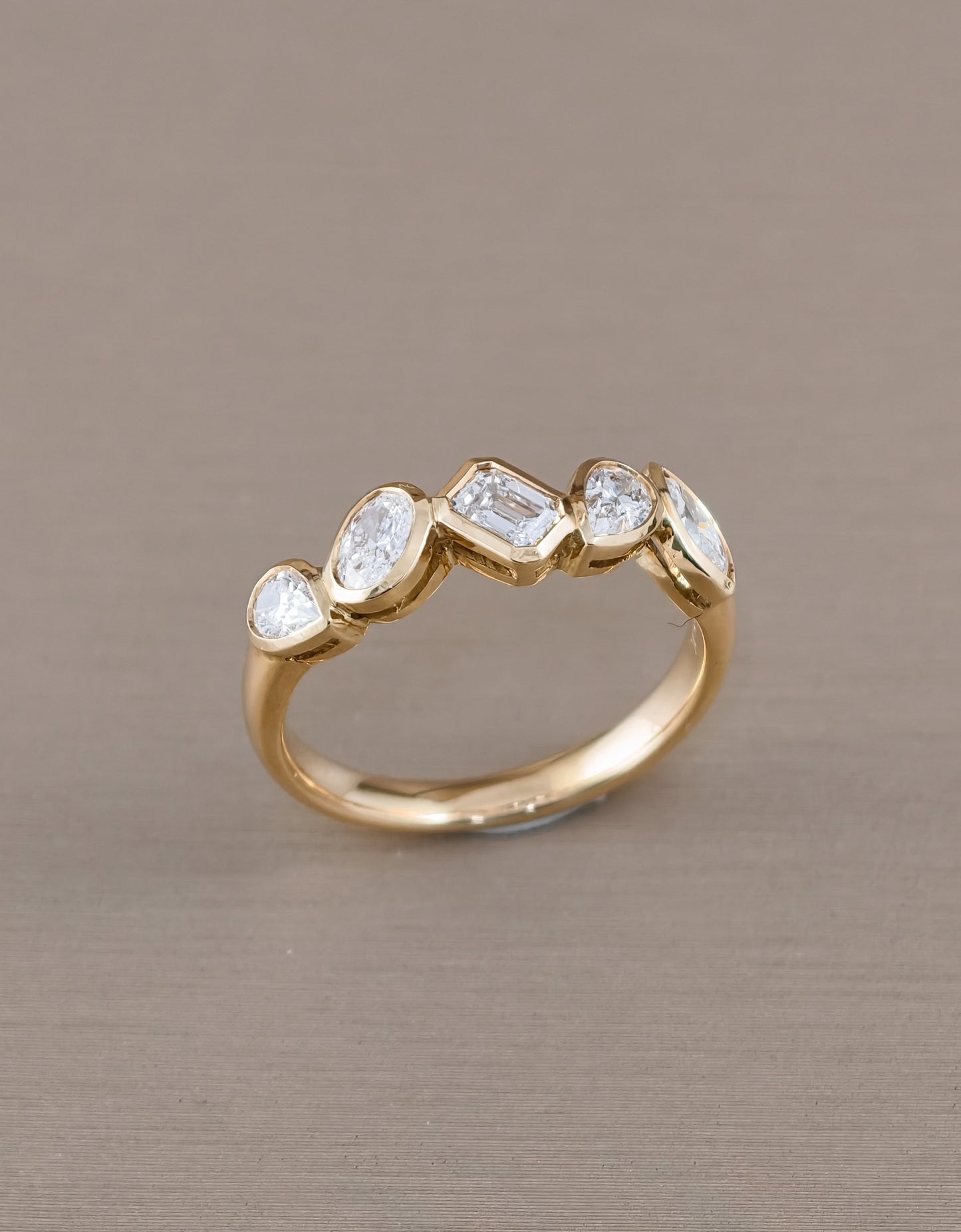 A gold ring featuring five clear gemstones in marquise, baguette, and oval shapes, displayed on a textured taupe background.