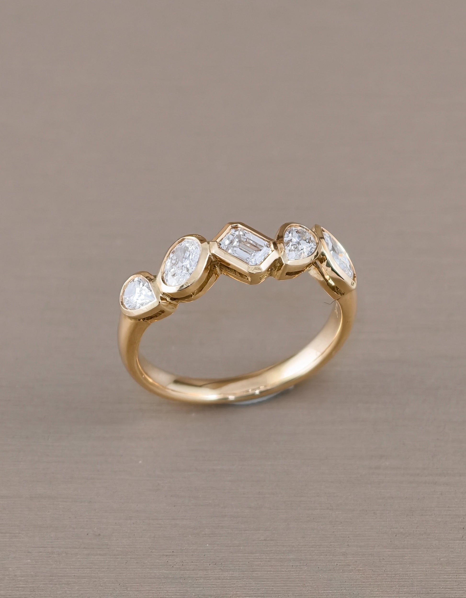 A gold ring featuring five clear gemstones in marquise, baguette, and oval shapes, displayed on a textured taupe background.