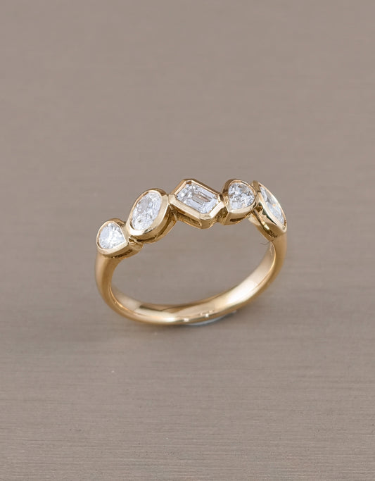 A gold ring featuring five clear gemstones in marquise, baguette, and oval shapes, displayed on a textured taupe background.